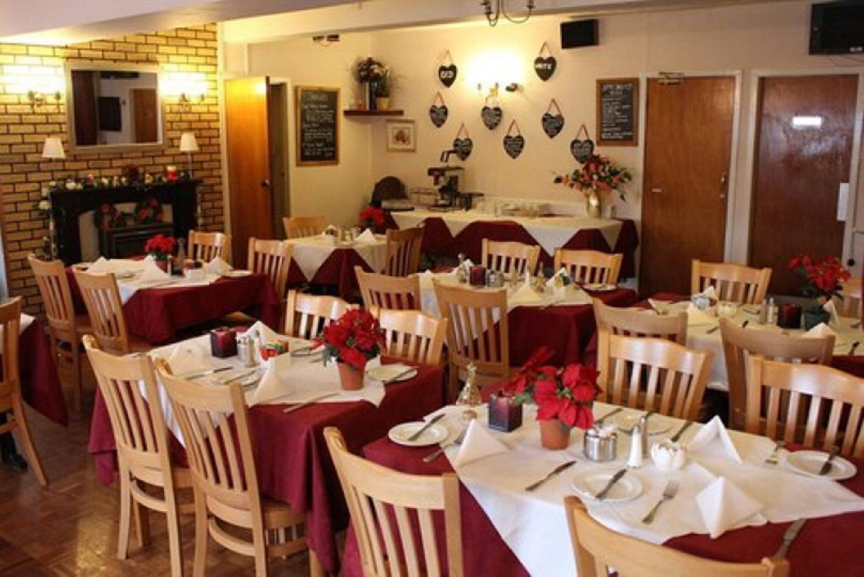 A restaurant with multiple tables, each set with white tablecloths and red napkins. There are wooden chairs around the tables, and a brick wall with a fireplace.