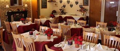 A restaurant with multiple tables, each set with white tablecloths and red napkins. There are wooden chairs around the tables, and a brick wall with a fireplace.
