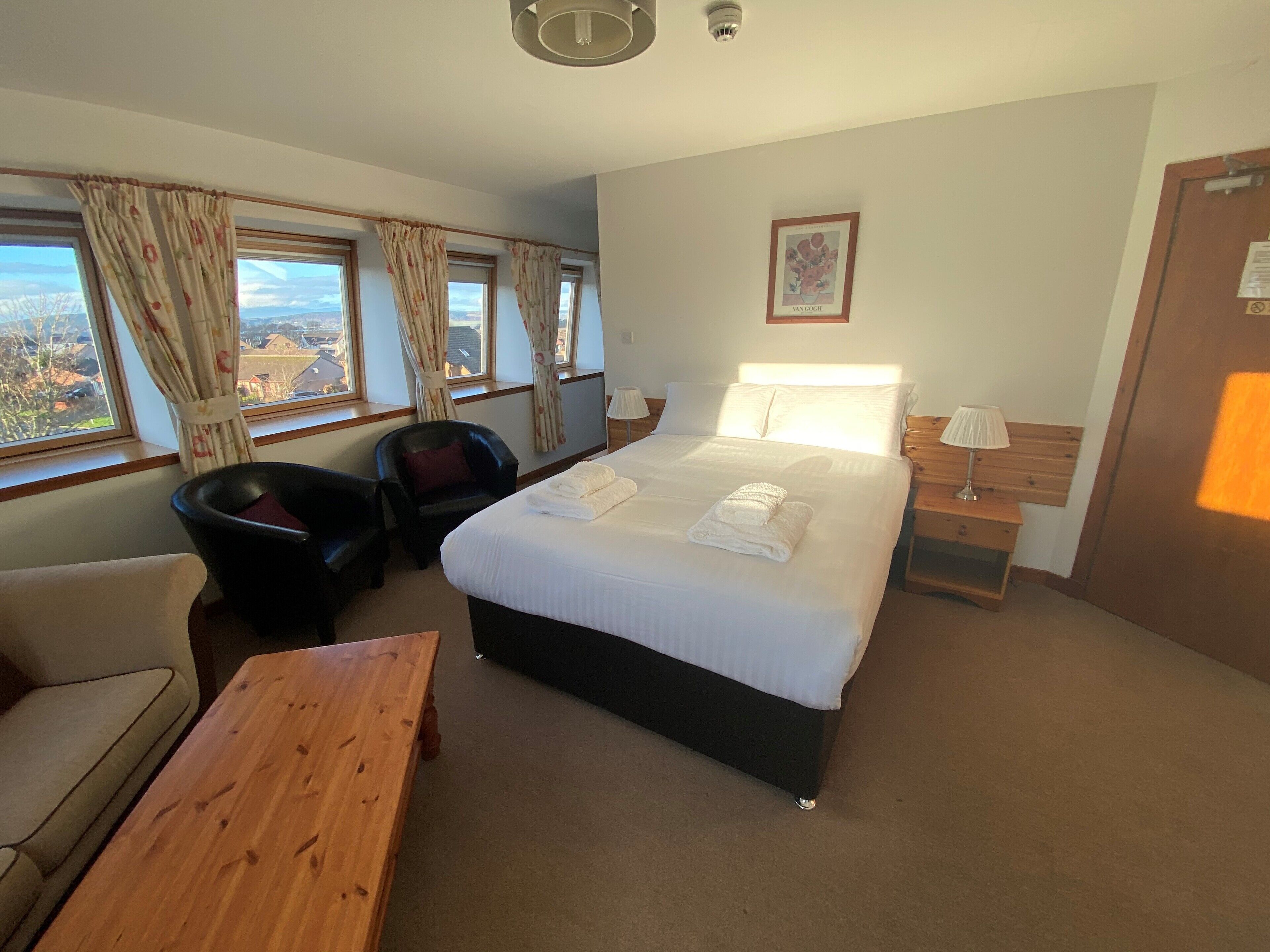 A hotel room with a bed, a chair, a wooden table, and a view of the outside through the windows.