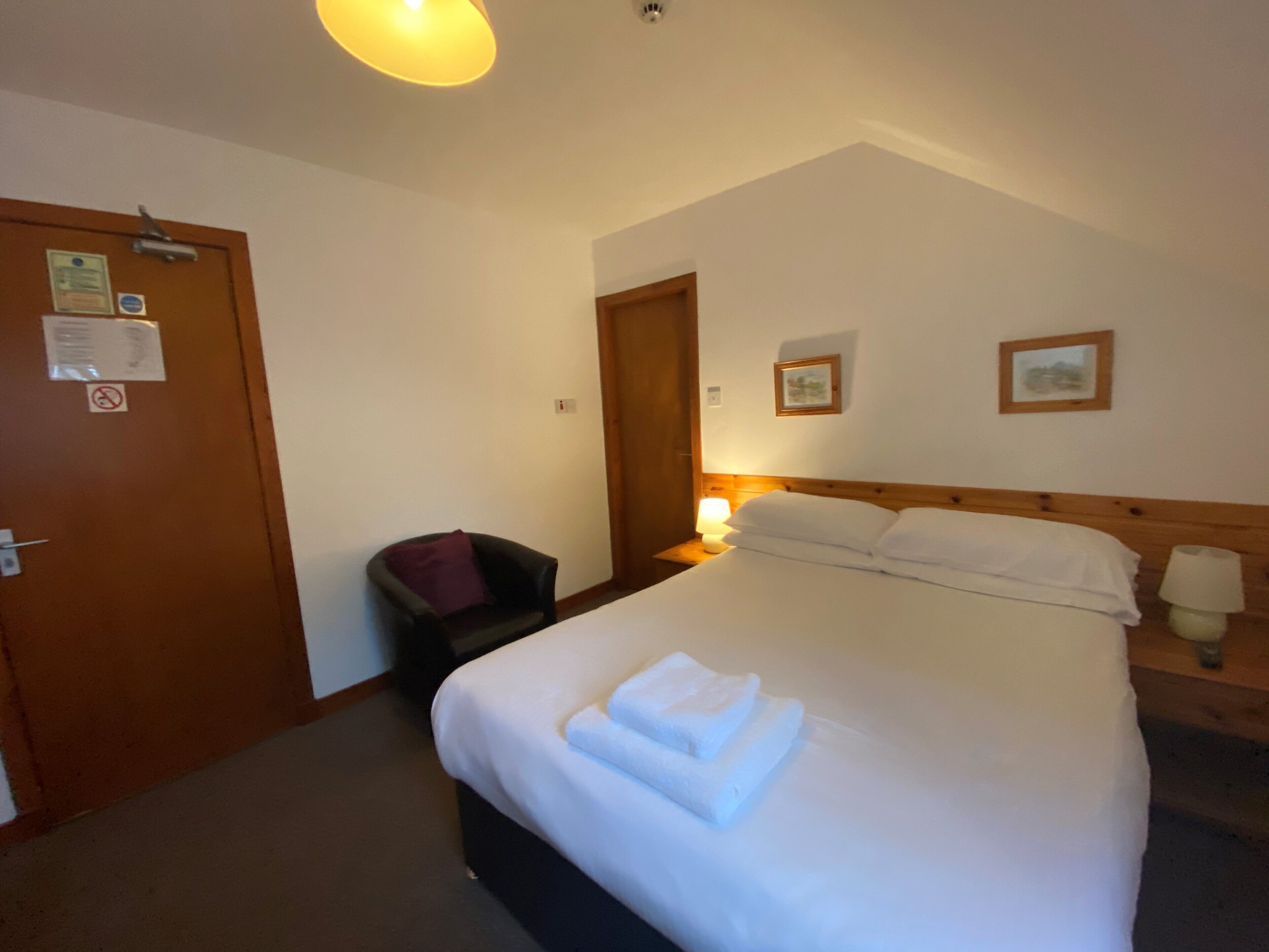 Et hotelværelse med en seng, en stol, et natbord med lampe og en dør med skilt.