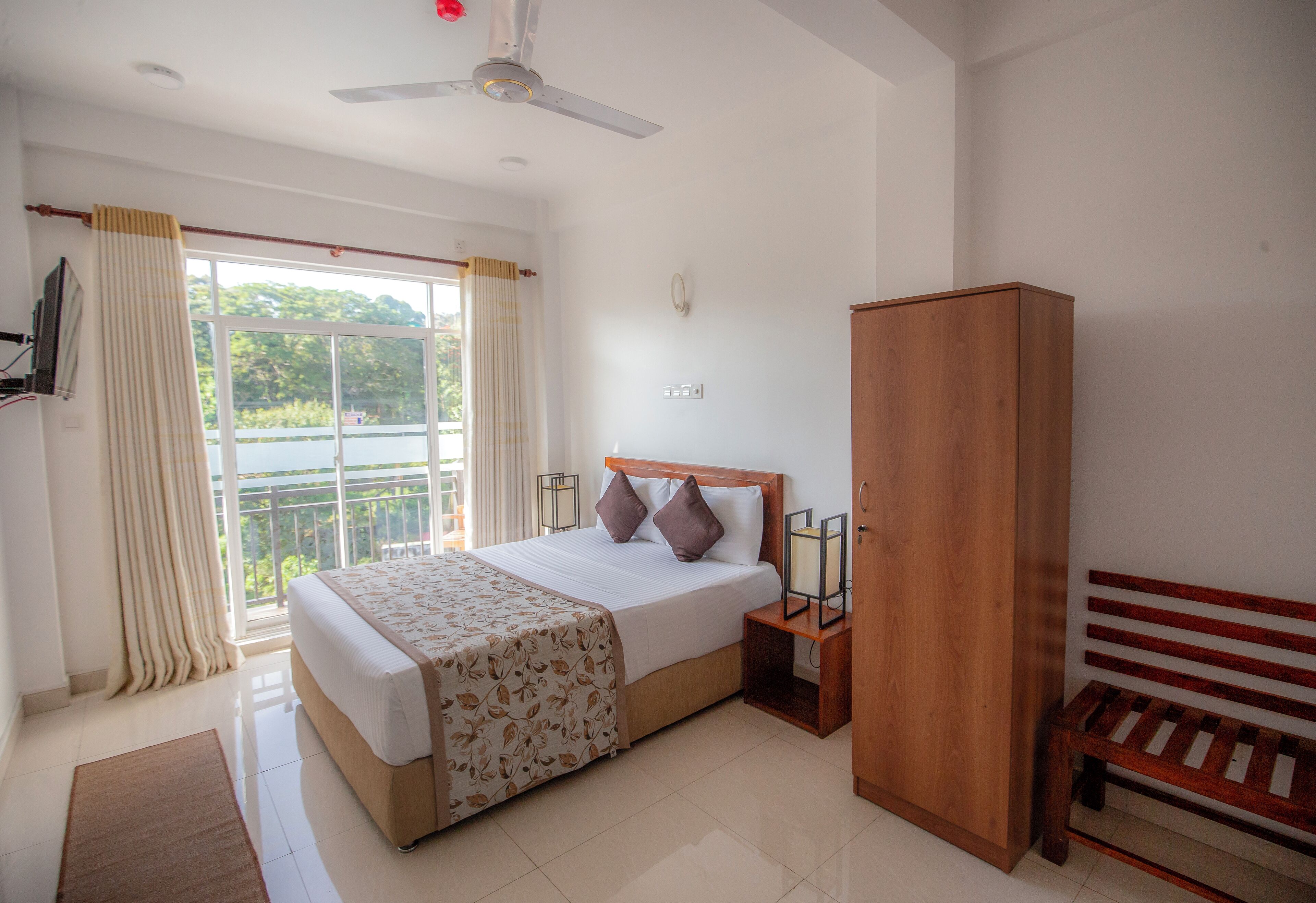 Une chambre d’hôtel avec un lit, une armoire en bois, un banc et un ventilateur de plafond.