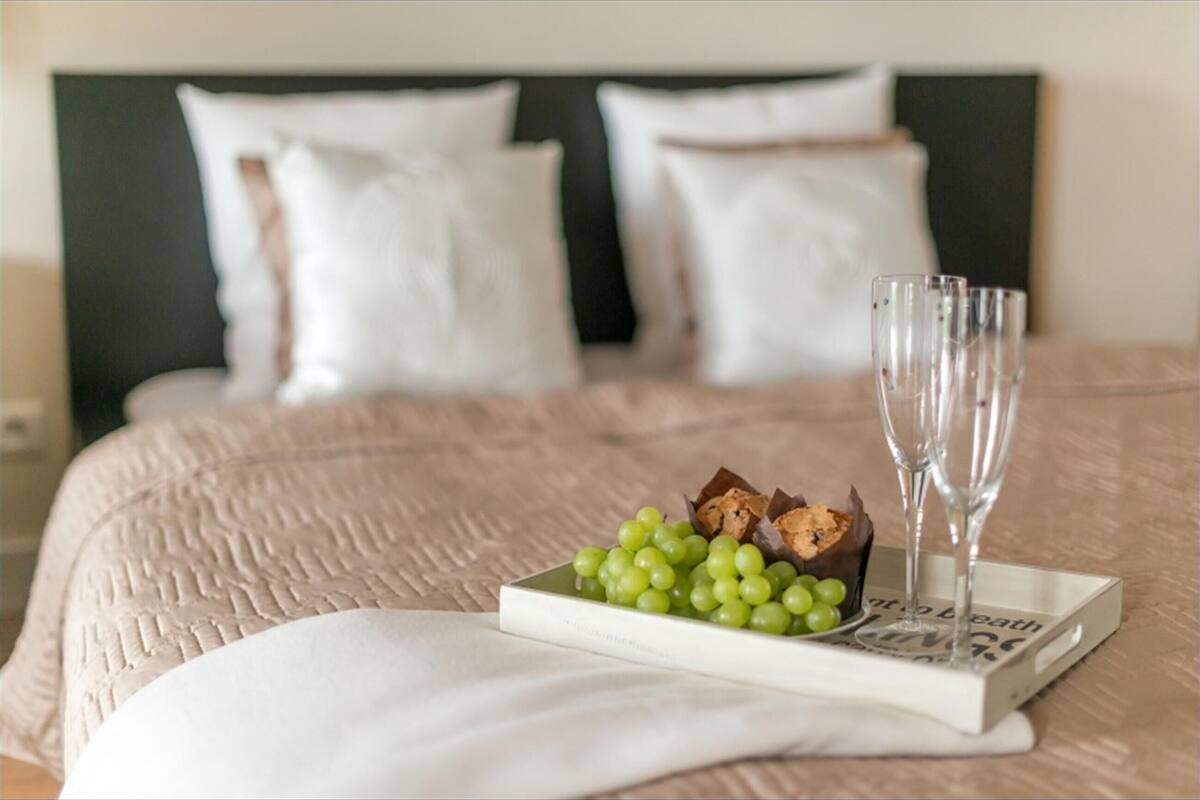 Una cama bien hecha con una colcha blanca y almohadas, una bandeja con uvas verdes y galletas de chocolate, y dos vasos vacíos en una bandeja junto a un libro titulado 'No se debe repetir'.