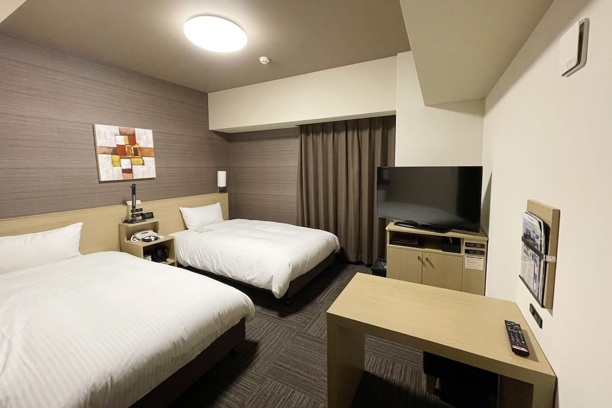 A hotel room with two beds, a desk, a television, and a ceiling light.
