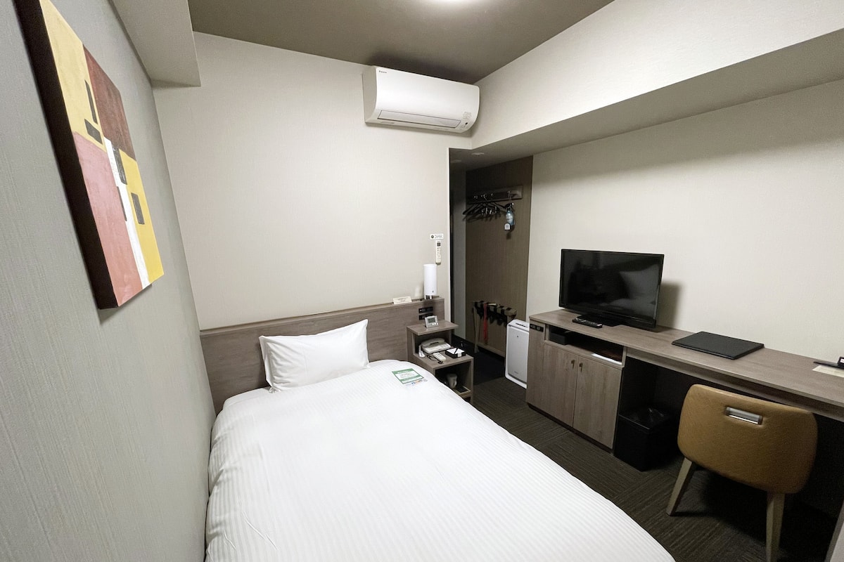 A hotel room with a bed, a desk with a television, a chair, a small refrigerator, and an air conditioning unit.