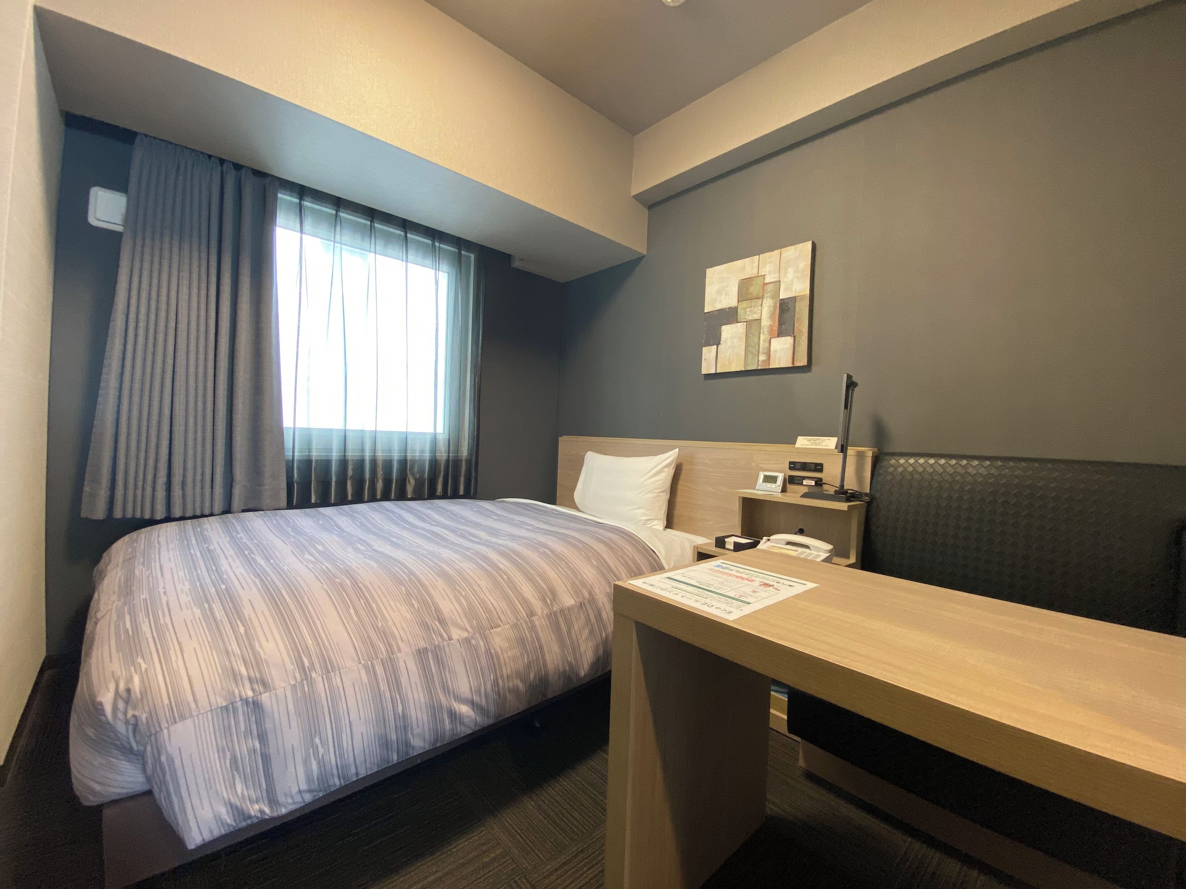Habitación de hotel con cama, escritorio, silla, mesita de noche, lámpara y ventana con cortinas.