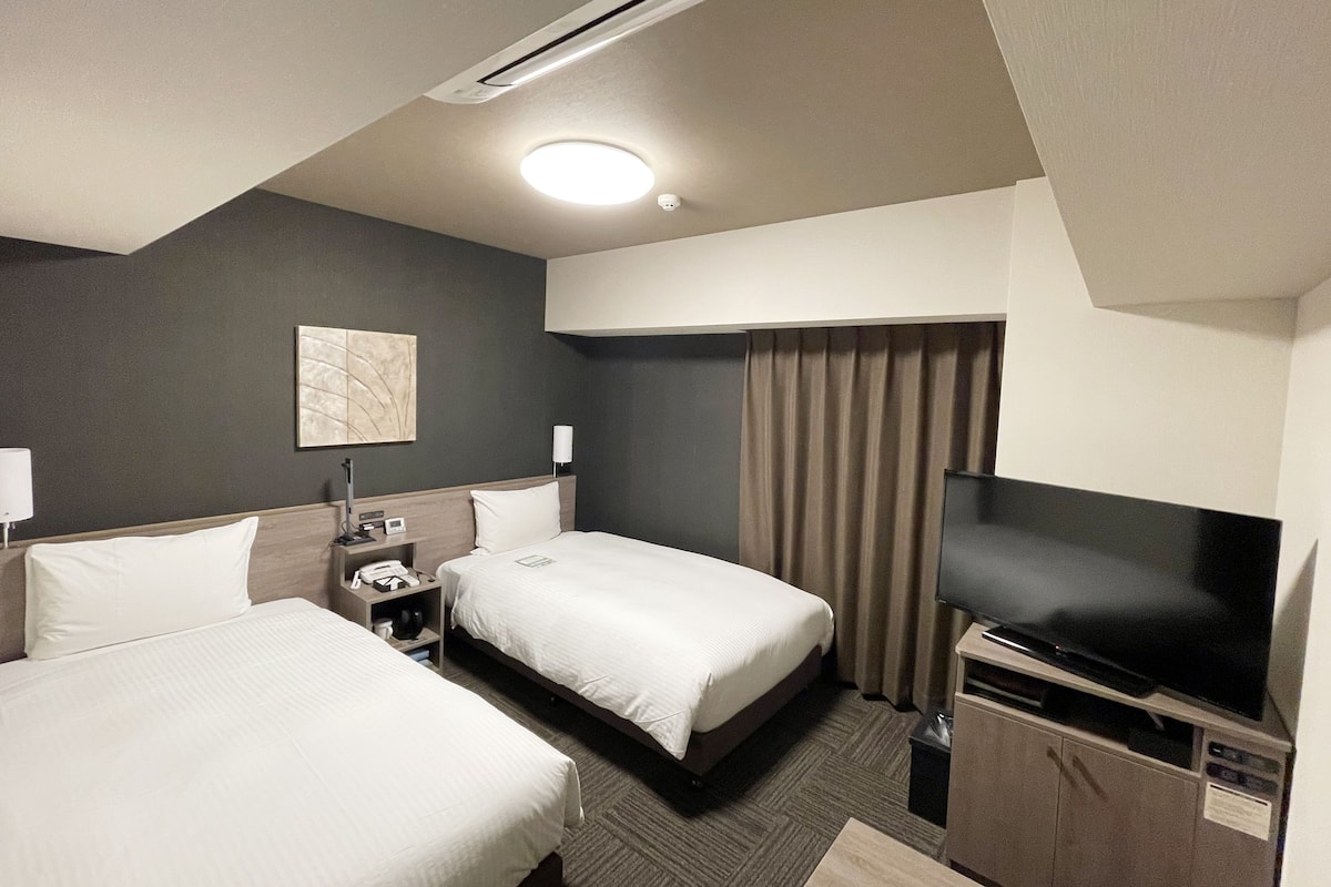 A hotel room with two beds, a television, a desk with a lamp, and a ceiling light.