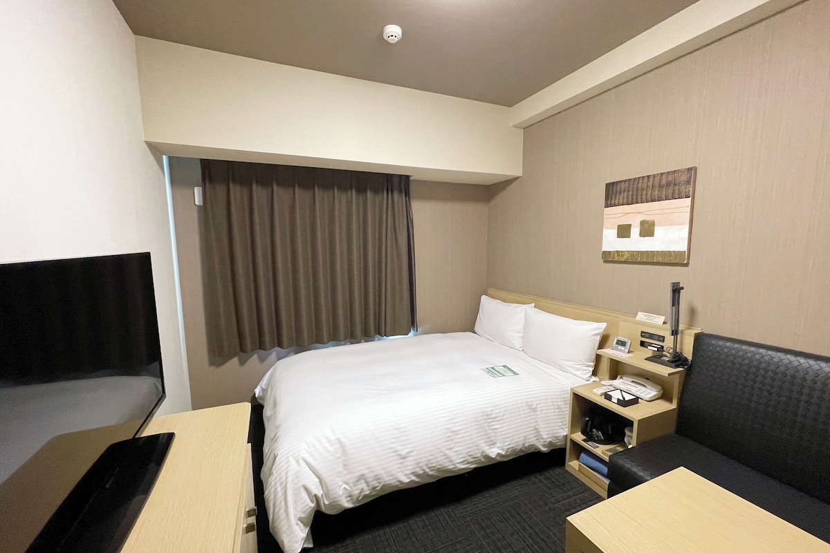 A hotel room with a bed, a desk, a chair, a nightstand, and a window with curtains.