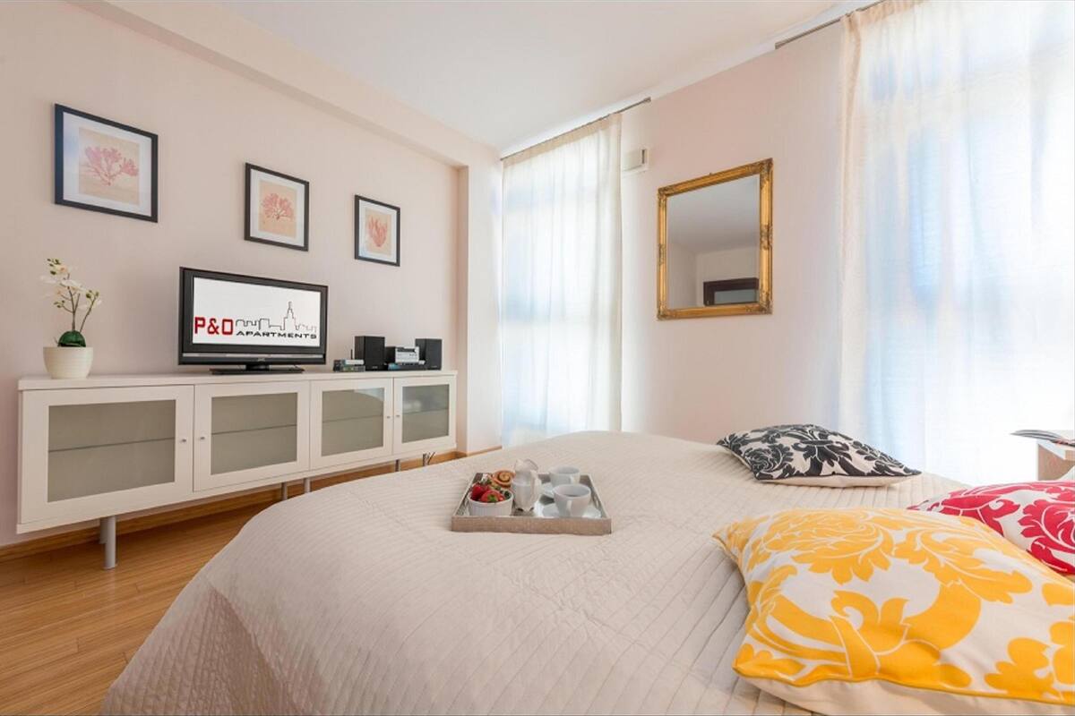 A modern hotel room with a bed, a TV on a cabinet, a tray with breakfast items, and framed pictures on the wall.