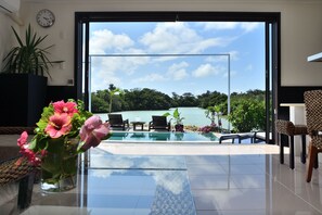 A modern room with a glass table, a vase of flowers, a view of a pool and a lake, and outdoor seating.