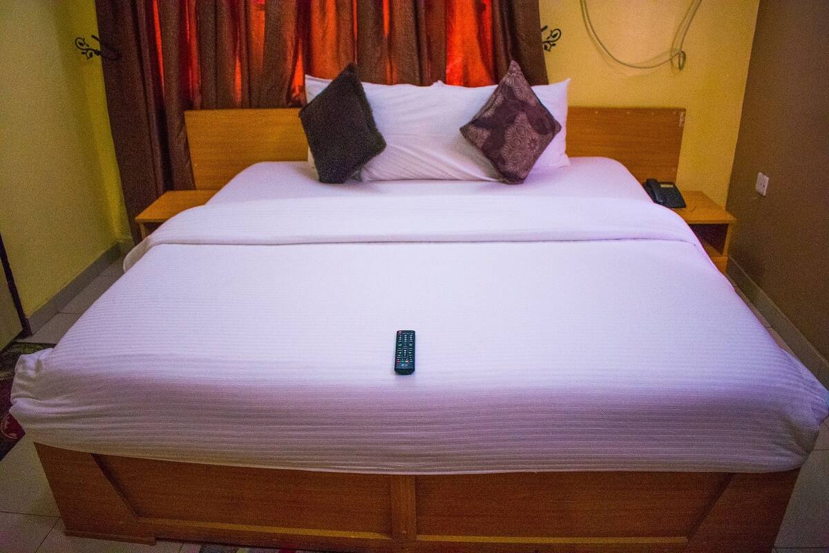 A neatly made bed with a remote control on it, a wooden headboard, and a nightstand with a phone.