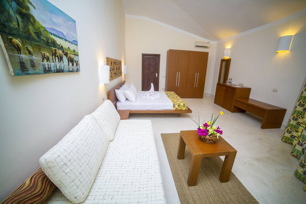 A modern, well-lit room with a sofa, a small table with flowers, a bed with a white sheet, wooden cabinets, and a painting on the wall.