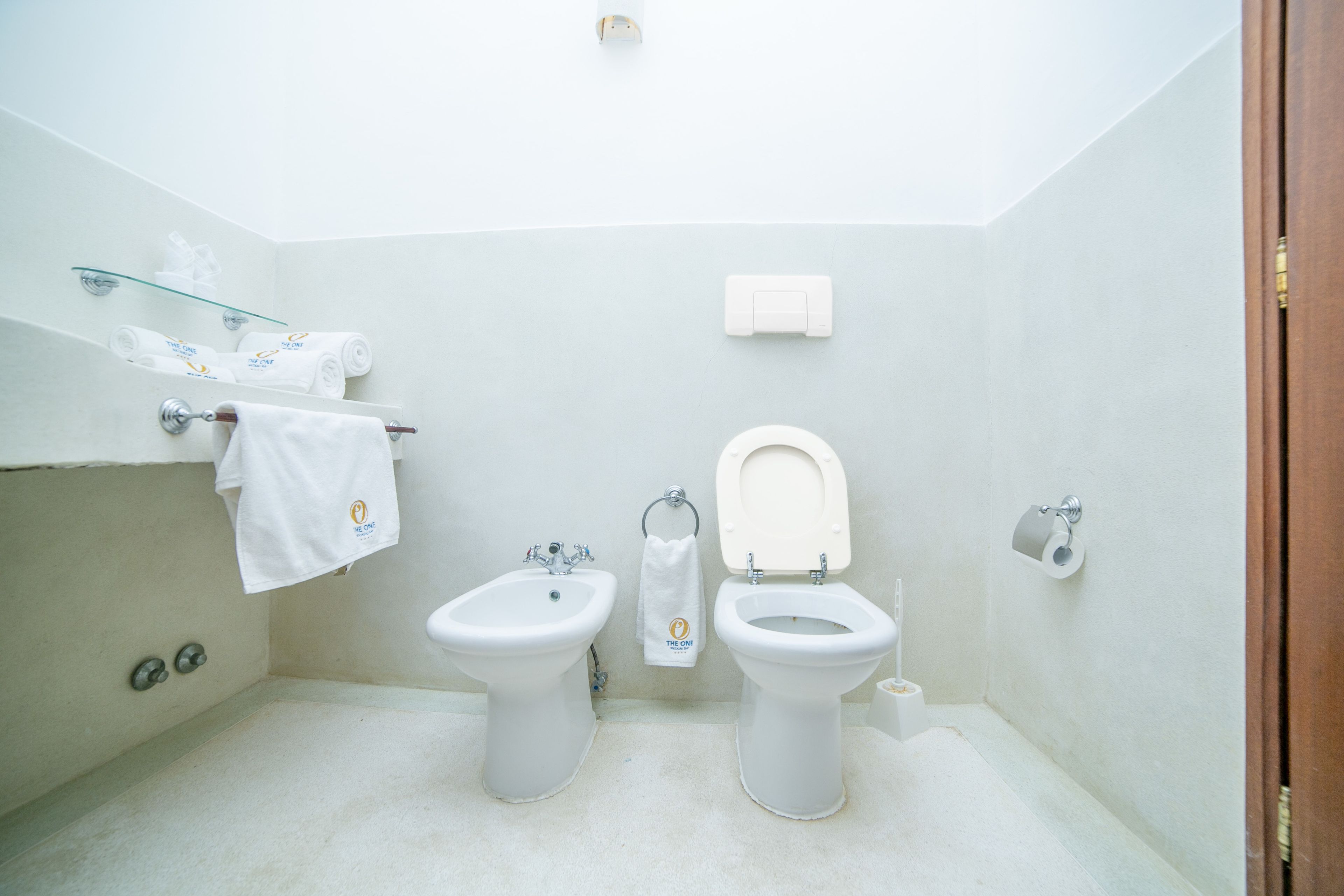 A bathroom with two toilets, a bidet, a glass shelf with towels, and a toilet paper holder.