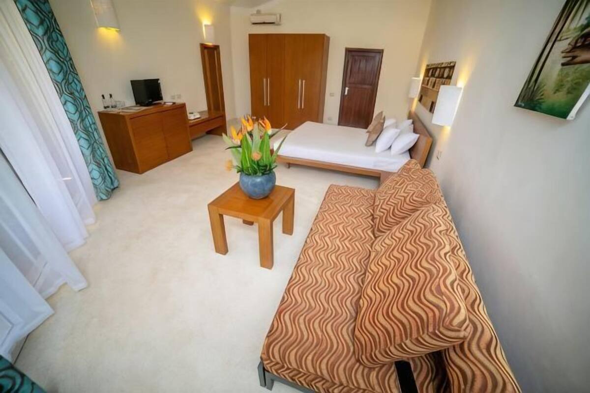 A compact room with a sofa, a small table with a vase, a bed, a wardrobe, and a television on a cabinet.