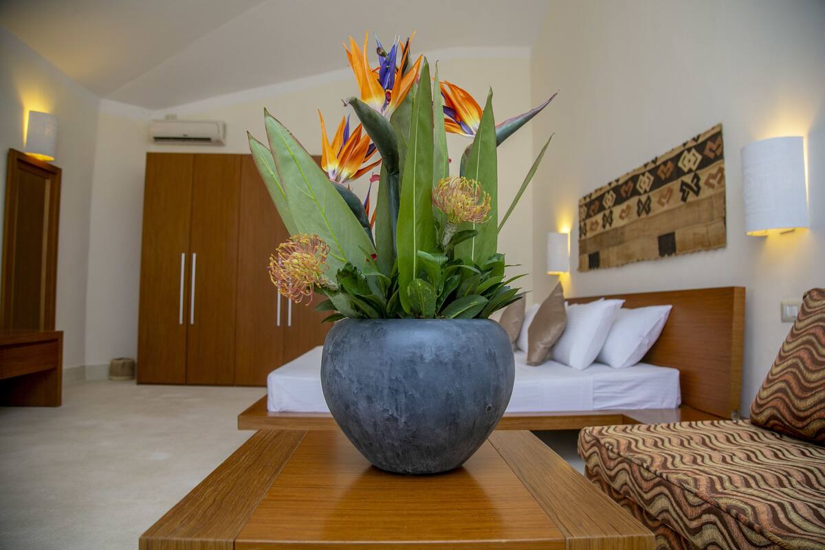 A modern hotel room with a large bed, a wooden nightstand, a potted plant with bird of paradise flowers, and a wall-mounted lamp.