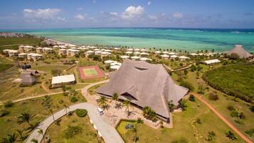 Eine Vogelperspektive zeigt ein Resort mit mehreren bungalowsähnlichen Gebäuden, einem zentralen Bauwerk mit einem Reetdach und freiem Blick auf den Ozean.
