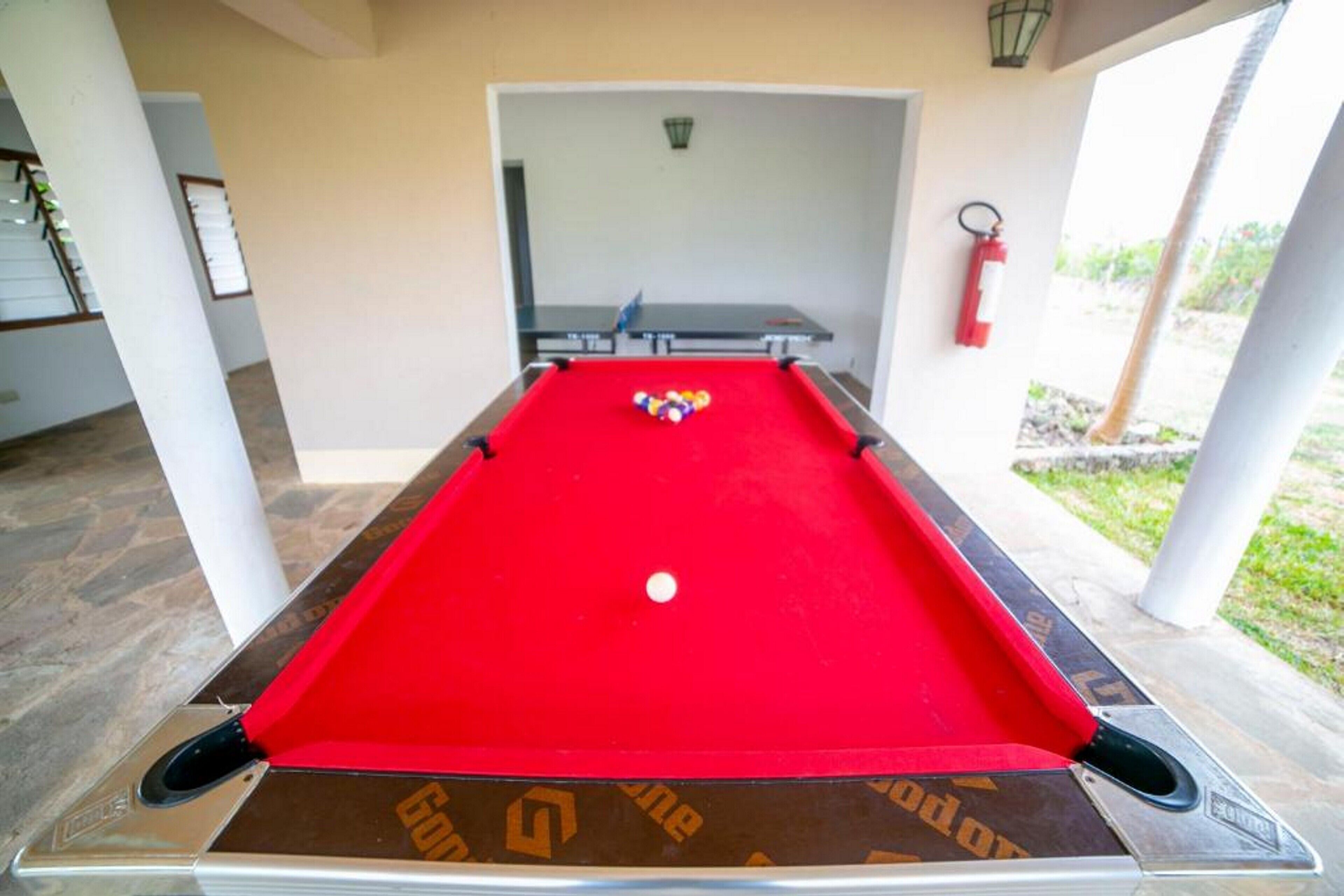 A pool table with a red felt surface and a white cue ball, surrounded by white pillars and a covered area with a view of greenery.