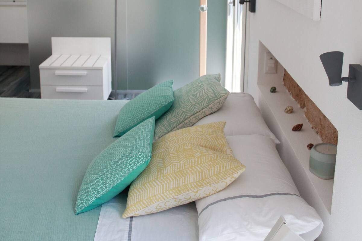 A bedroom with a bed, pillows, a bedside table, a nightstand, a mirror, and a wall-mounted artwork.