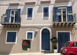 Un bâtiment avec des volets bleus, une porte d’entrée centrale en arc de cercle et des balcons dotés de balustrades.