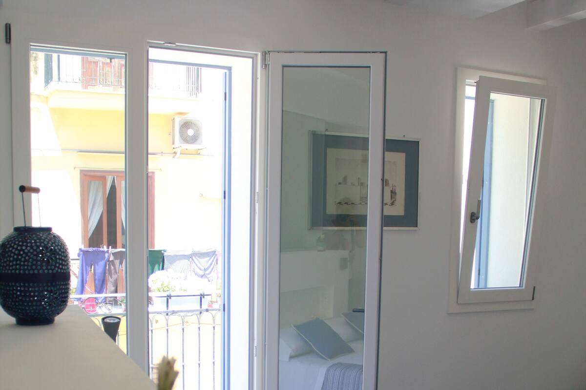 A room with a bed, a small table with a potted plant, and a sliding glass door leading to an outdoor area.