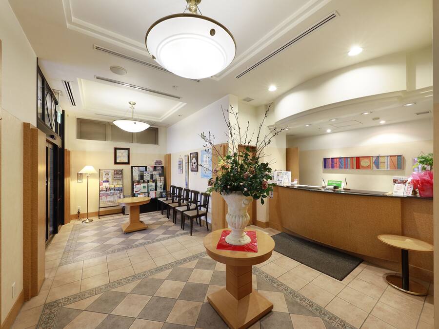 Un espace d’accueil comprenant un comptoir d’enregistrement, un coin salon avec des chaises, un vase décoratif avec des fleurs et un sol carrelé.