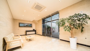 A modern lobby with a beige tiled floor, a wooden sofa, a small table, and a large potted tree. - Run Hotel (Seogwipo)