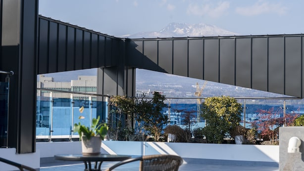 A modern building with a glass facade, a mountain view, and outdoor seating with potted plants. - Run Hotel (Seogwipo)