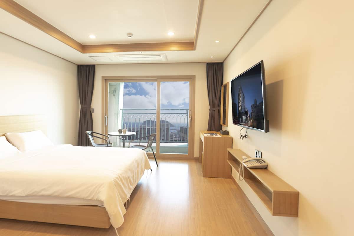 A hotel room with a bed, a TV, a desk, and a balcony with a city view.