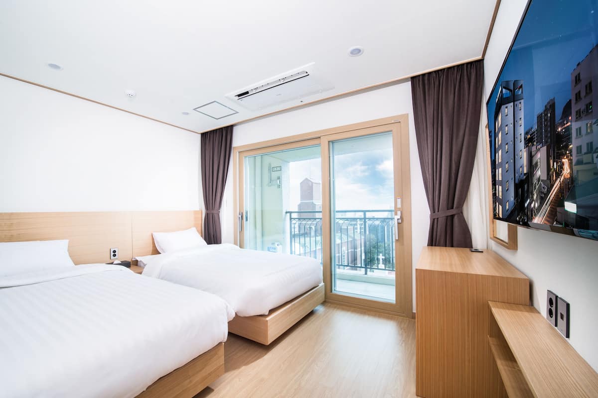 A hotel room with two beds, a large window with a view of the city, a TV, and a wooden desk.