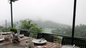 A wooden table set for dining with plates, cutlery, and glasses, overlooking a river and greenery. - Nest and Hives Mukteshwar (Dhari)