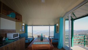 A dining area with a table and chairs, a microwave, and a view of the mountains through large windows. - Nest and Hives Mukteshwar (Dhari)