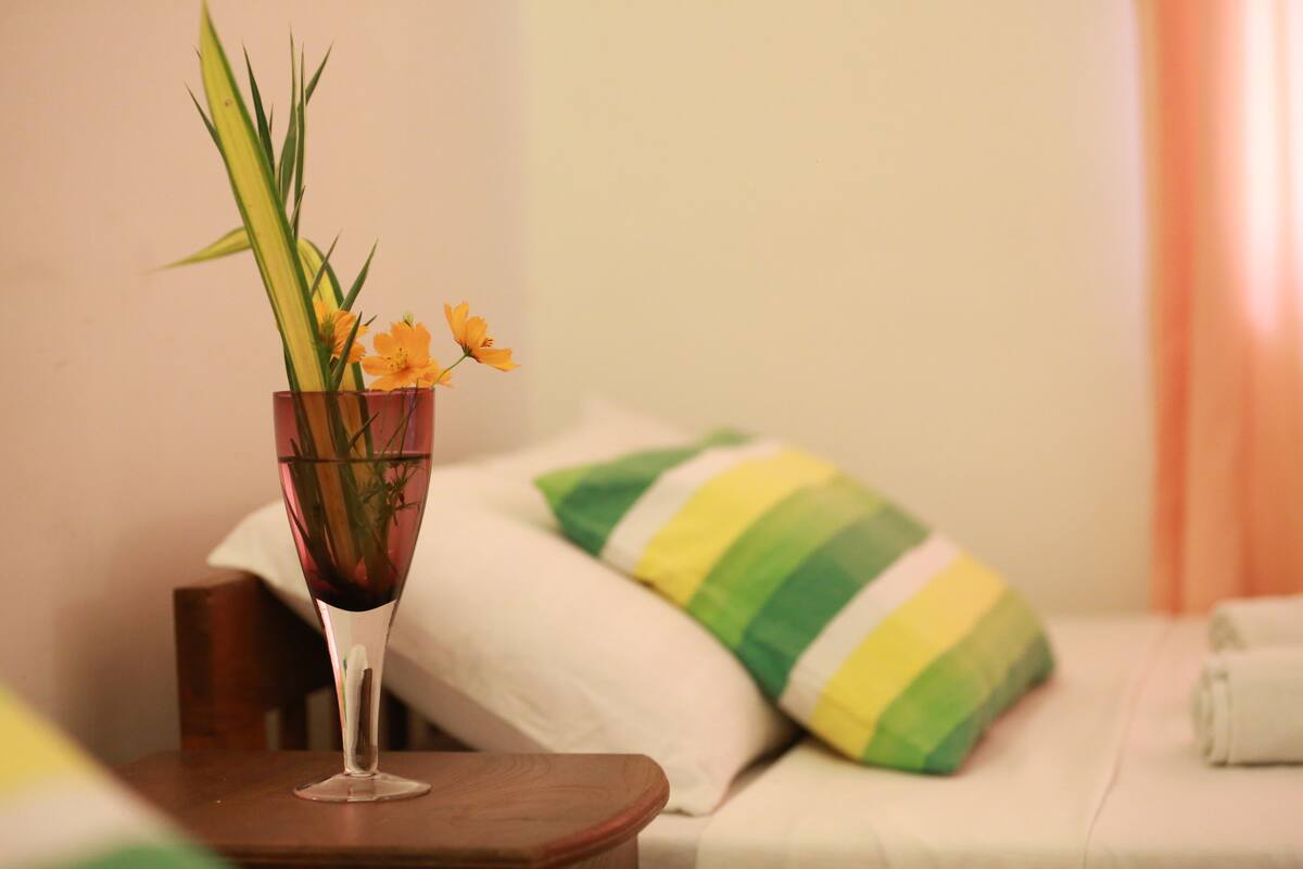 Quarto com cama, travesseiro listrado em verde e amarelo, um vaso com flores e uma mesa de madeira.
