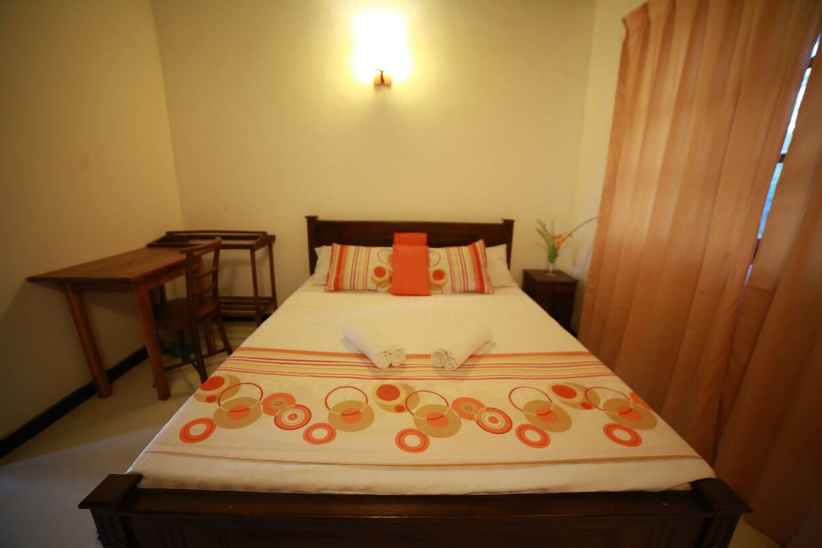 Uma cama bem arrumada com colcha listrada, uma mesinha de cabeceira de madeira com um vaso e uma escrivaninha de madeira com cadeiras em um quarto com paredes bege.