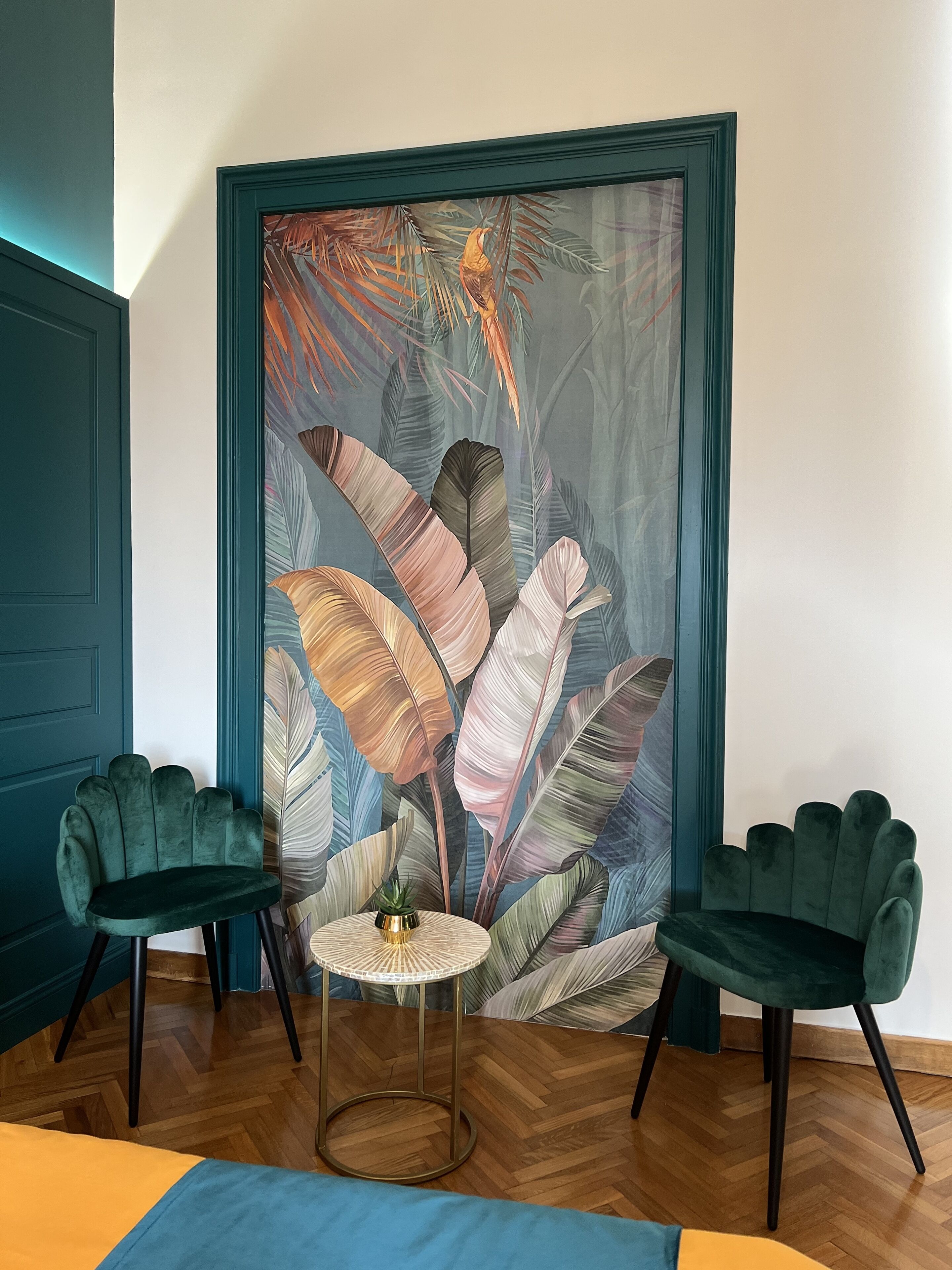 A room with a large tropical leaf painting, two green armchairs, a small round table with a plant, and a herringbone wooden floor.