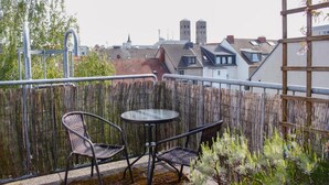 A balcony with two chairs and a small table, surrounded by a bamboo fence, overlooking a cityscape with traditional buildings and a church steeple. - Hotel Pilar Garni (Cologne)