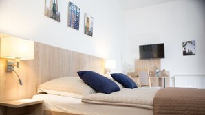 A hotel room with a bed, bedside table, lamp, TV, and artwork on the wall. - Hotel Pilar Garni (Cologne)
