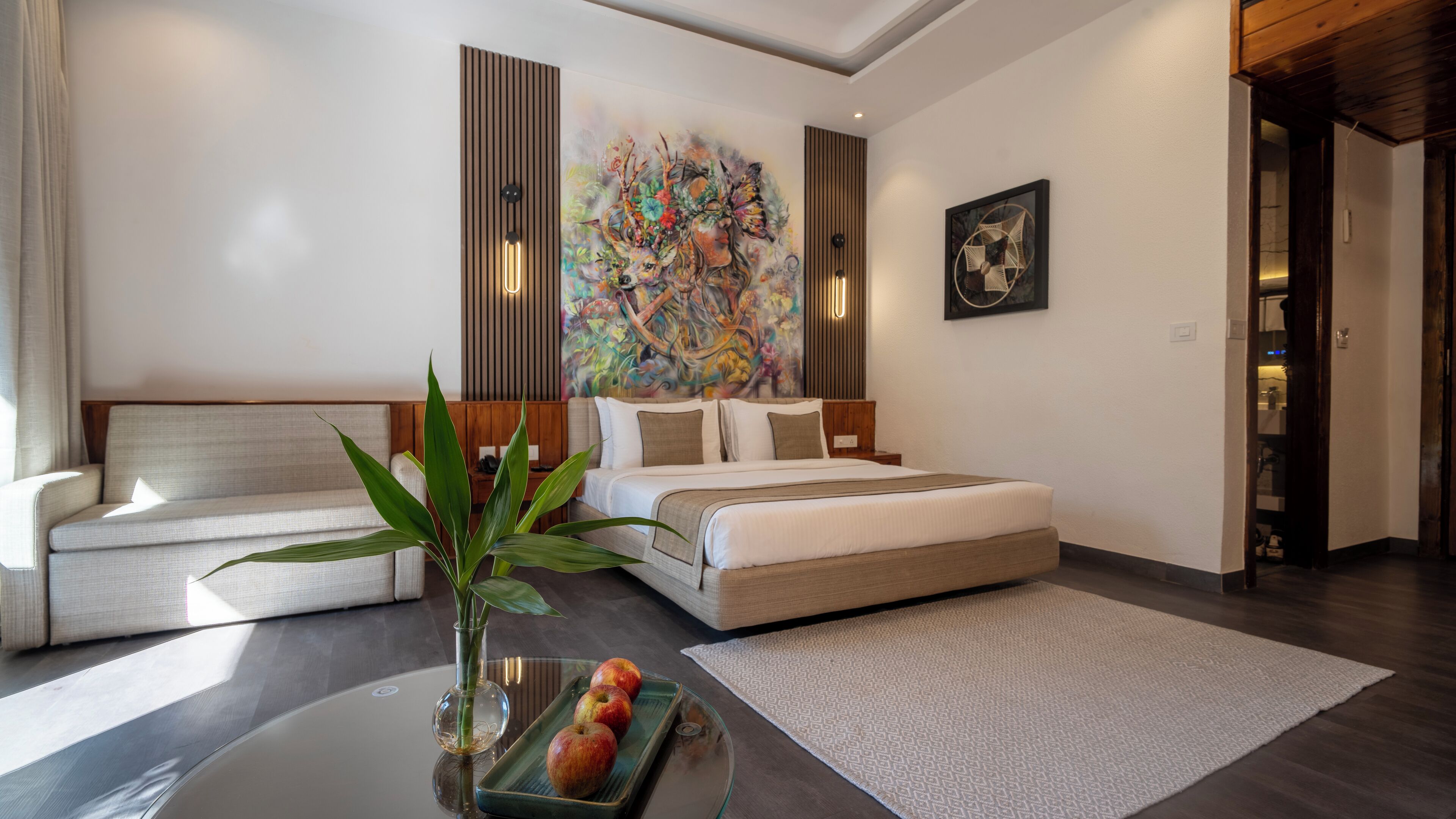 A modern hotel room with a bed, a sofa, a dining table with apples, and a large abstract painting on the wall.