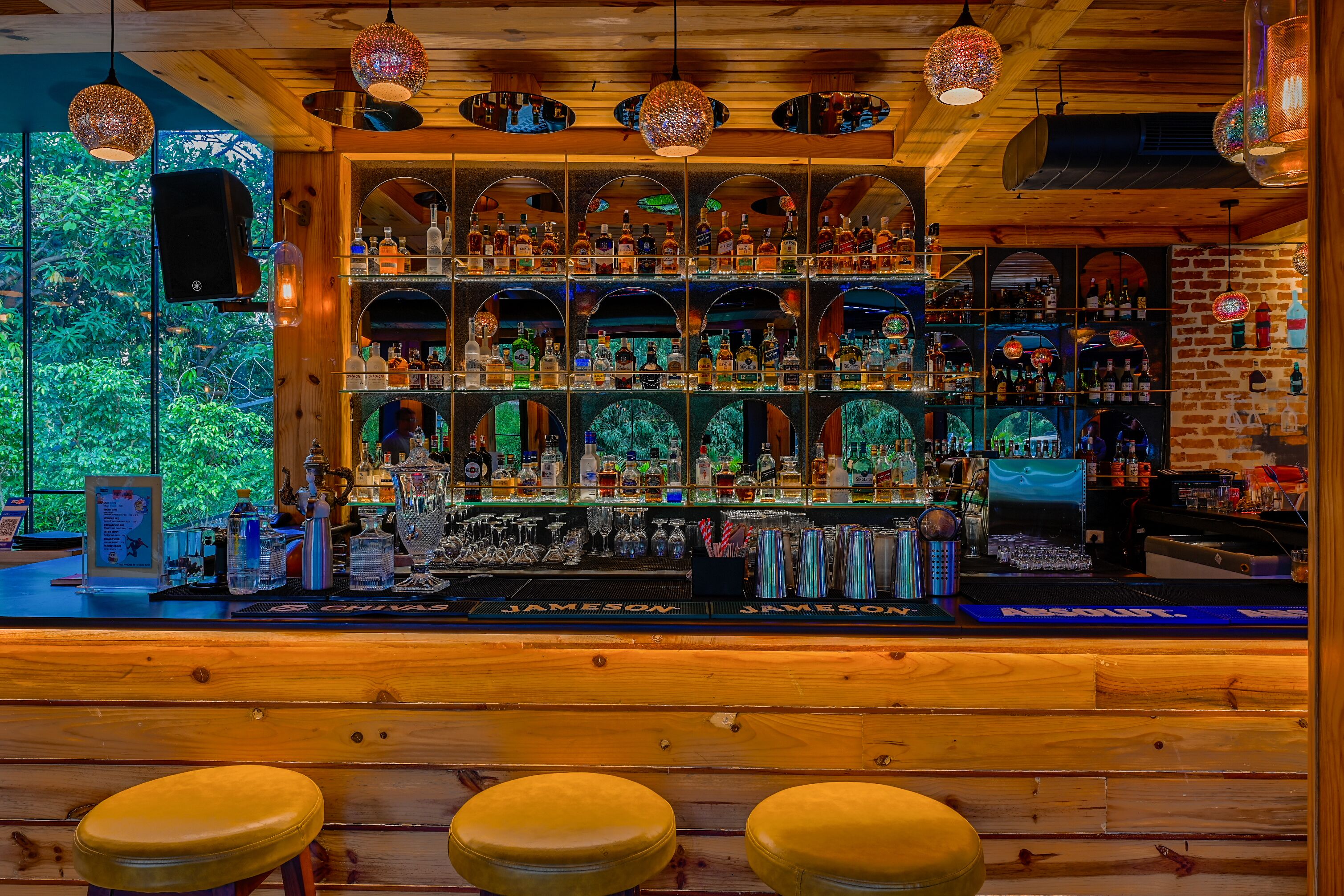 A bar with wooden shelves stocked with various bottles and glasses, a wooden counter, and three stools in the foreground.