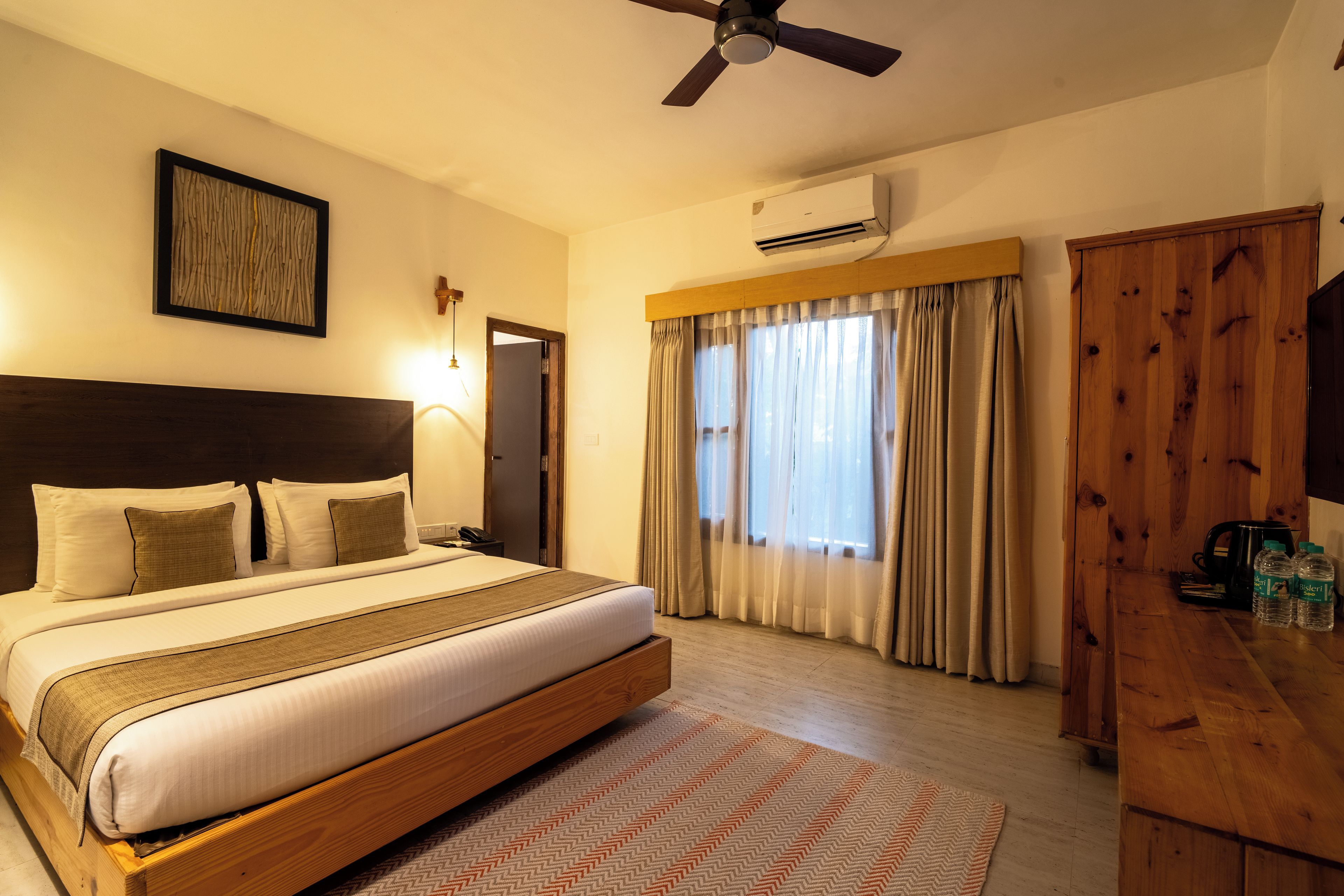 A hotel room with a large bed, a ceiling fan, an air conditioning unit, a wooden wardrobe, and a window with curtains.