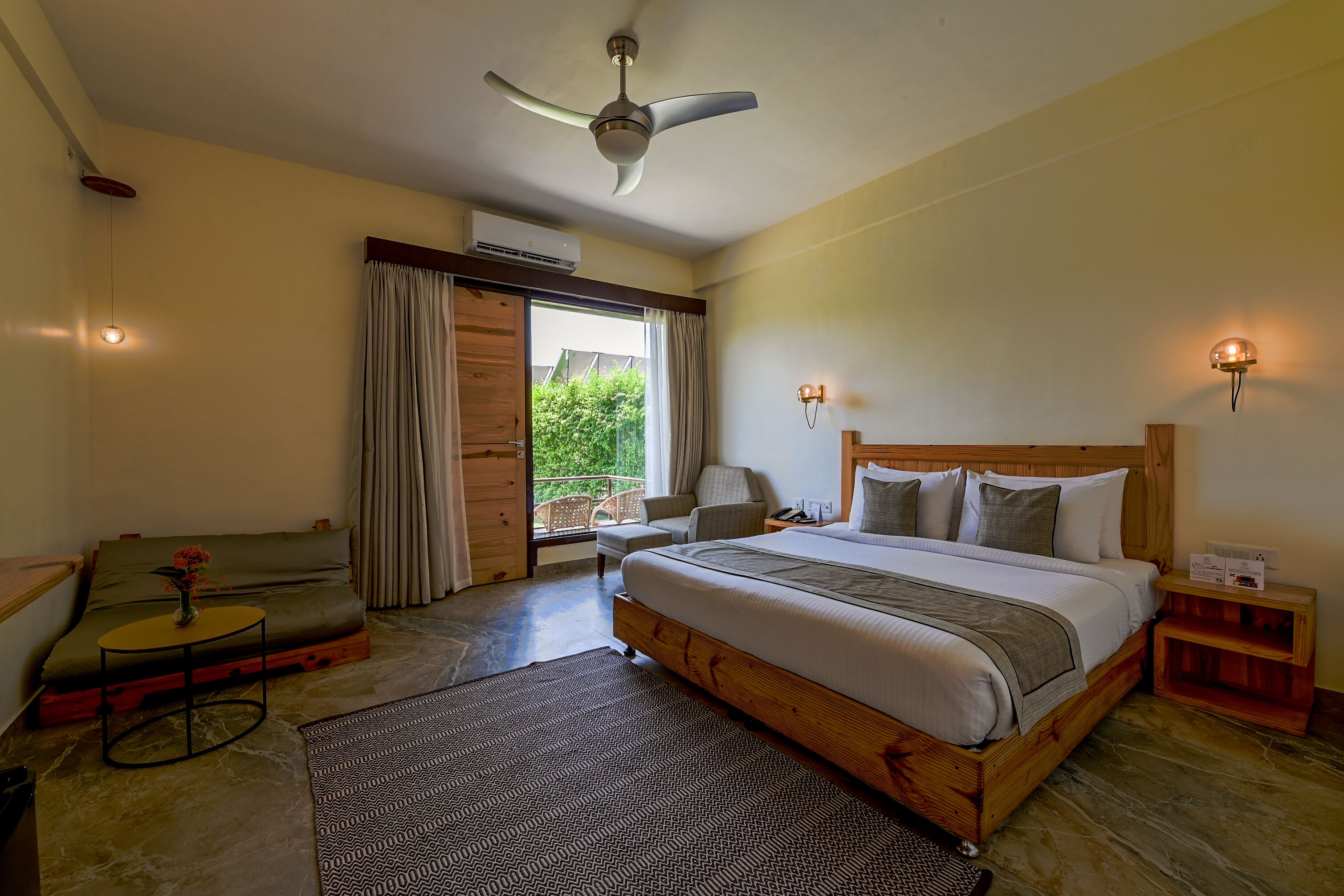A bedroom with a large bed, a ceiling fan, a window with curtains, a small table with a vase, and a nightstand with a lamp.