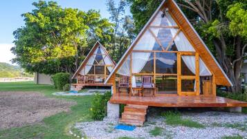 Duas cabanas de madeira triangulares com paredes de vidro, área de estar ao ar livre e gramado com árvores.
