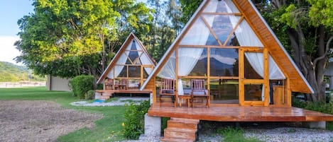 Due cabine di legno triangolari con pareti in vetro, mobili da esterno e una zona erbosa con alberi.