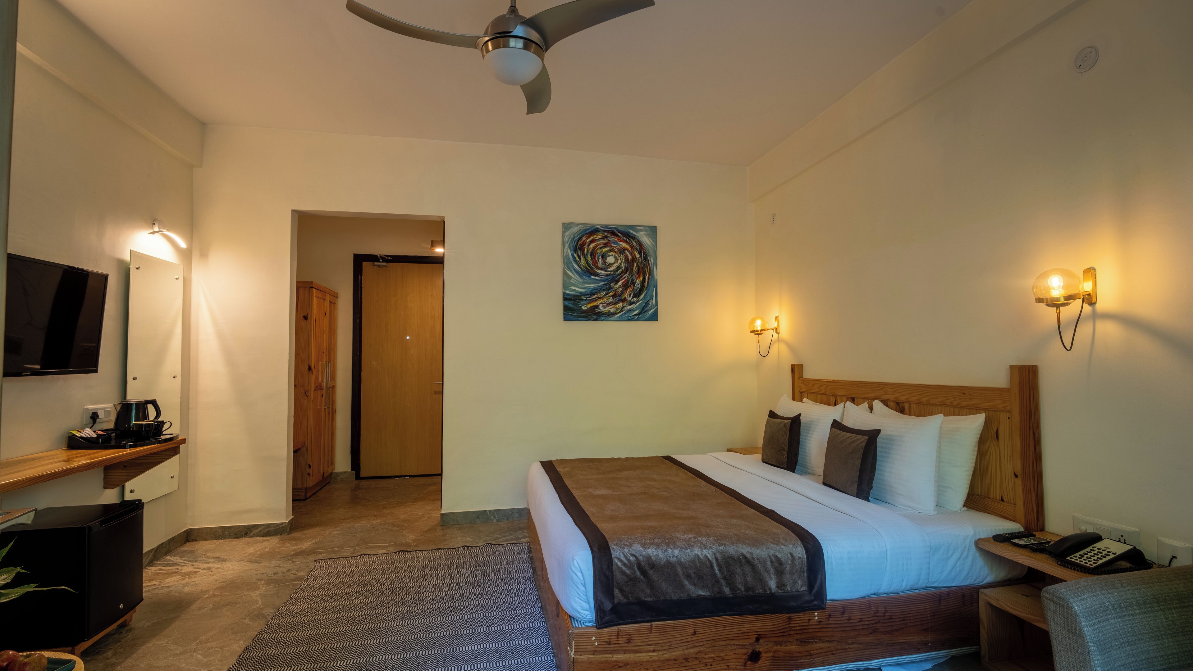 A hotel room with a bed, bedside table, lamp, TV, and a painting on the wall.