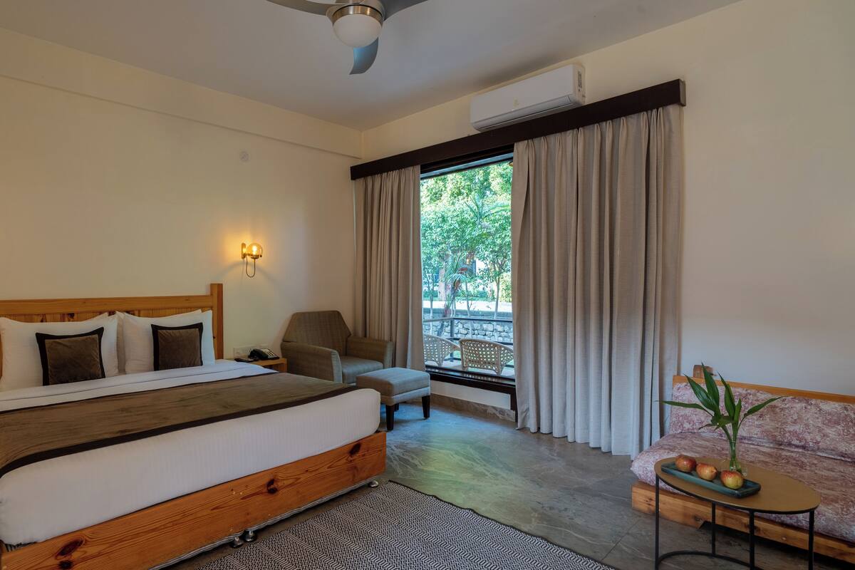 A bedroom with a wooden bed, two bedside lamps, a chair, a small table with fruit, and a view of greenery outside.