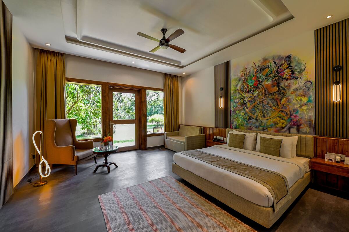 A modern hotel room with a large bed, a ceiling fan, a window with curtains, a painting on the wall, and a small table with a lamp.