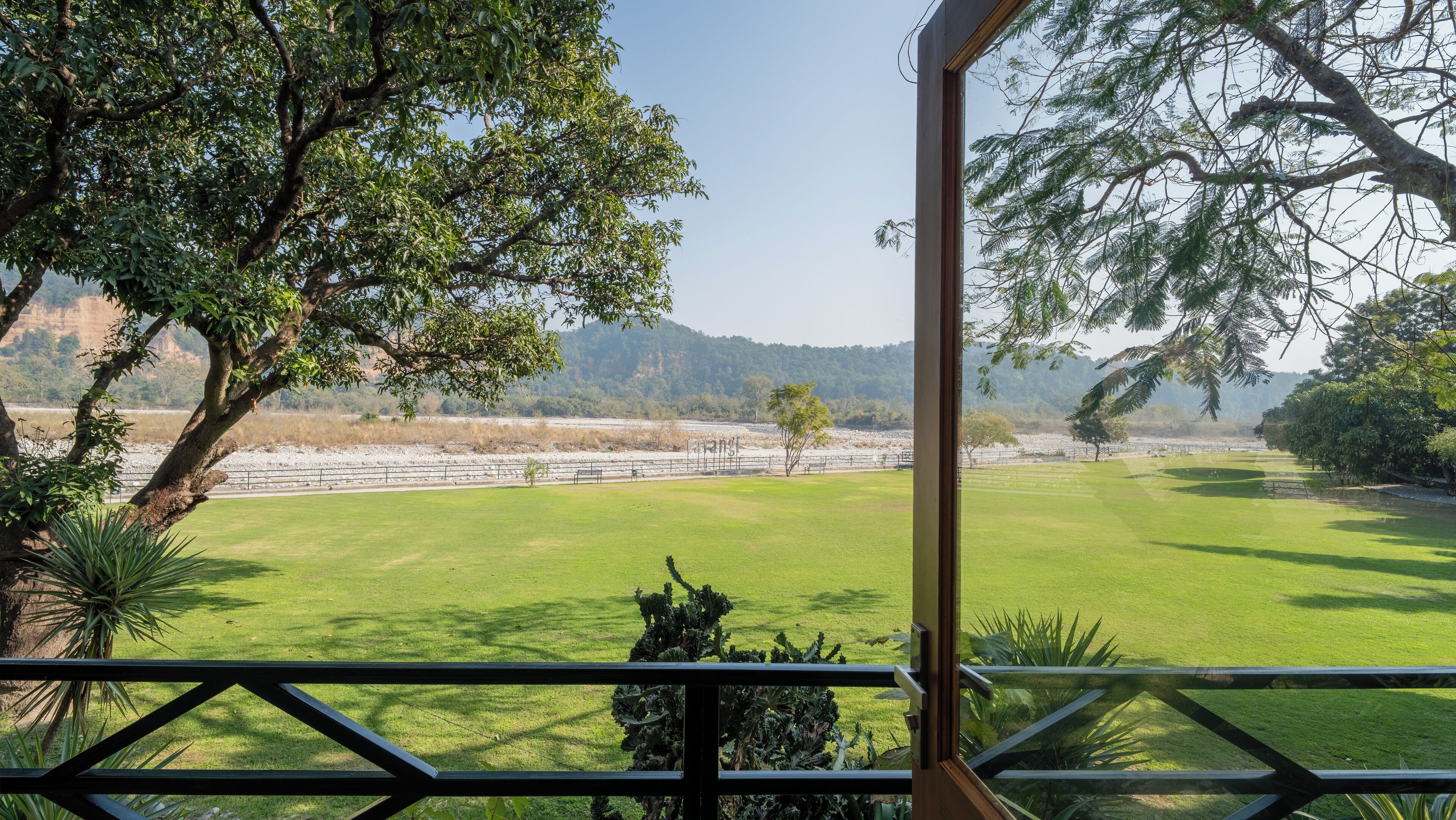 A view from a balcony overlooking a well-maintained lawn with a fence, trees, and a distant hilly landscape.