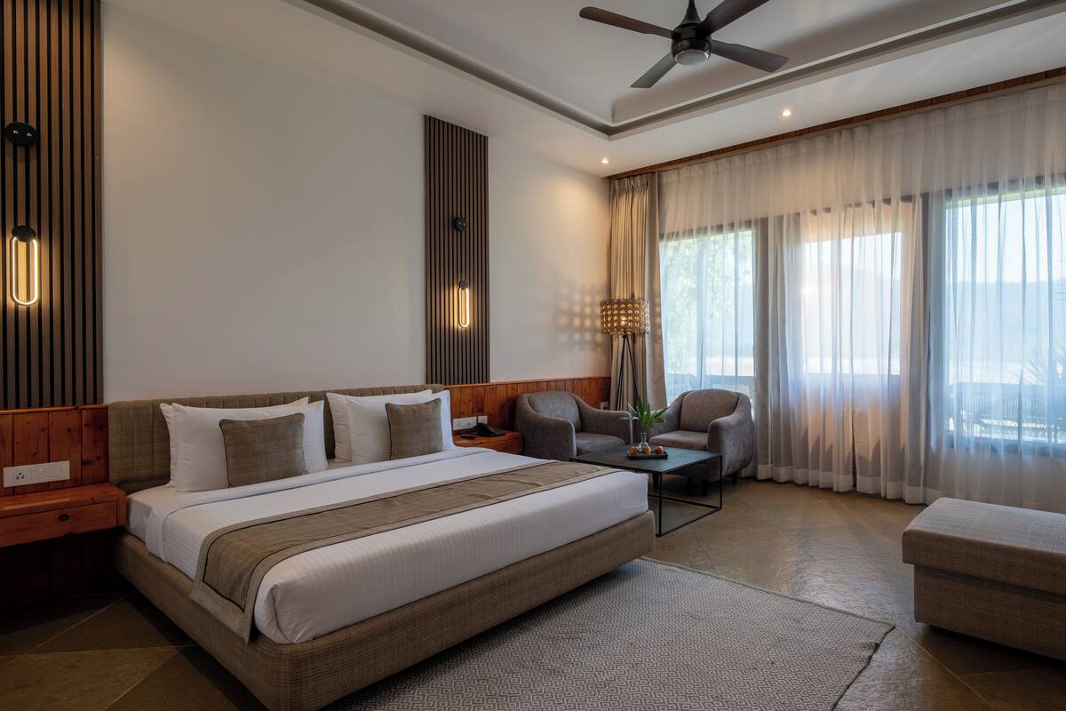 A modern hotel room with a large bed, two armchairs, a desk, and a ceiling fan.