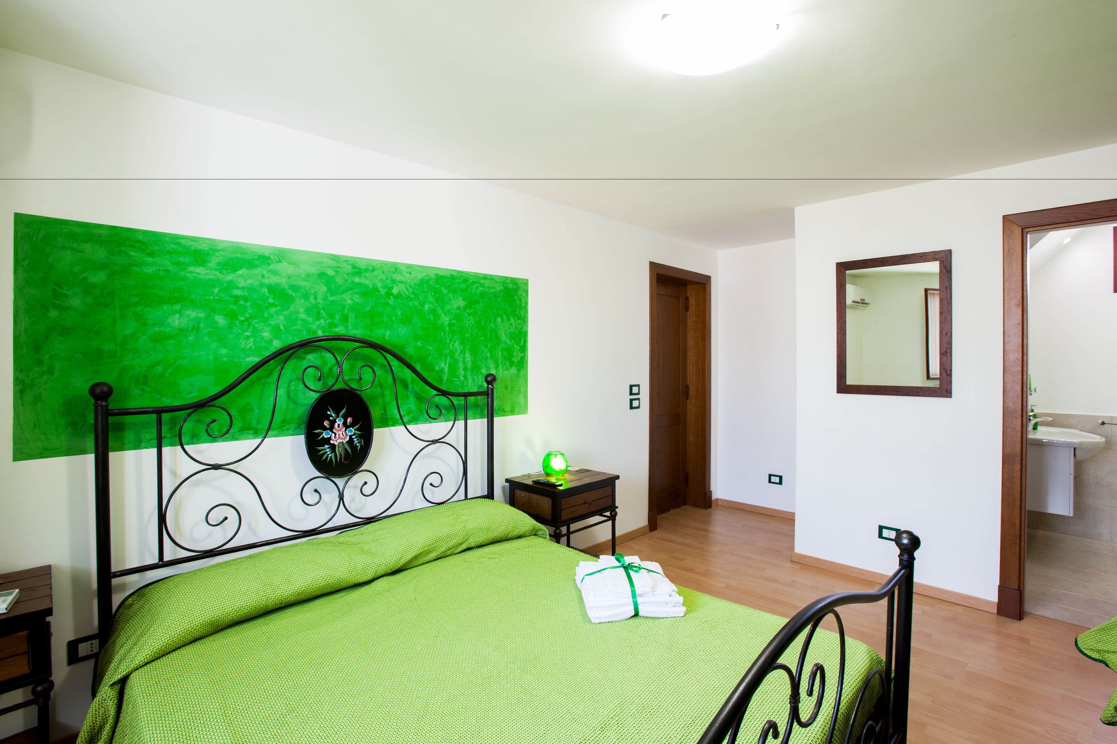 Un dormitorio con colcha verde, pared verde, suelo de madera, mesita de noche con lámpara y un baño visible a través de una puerta abierta.