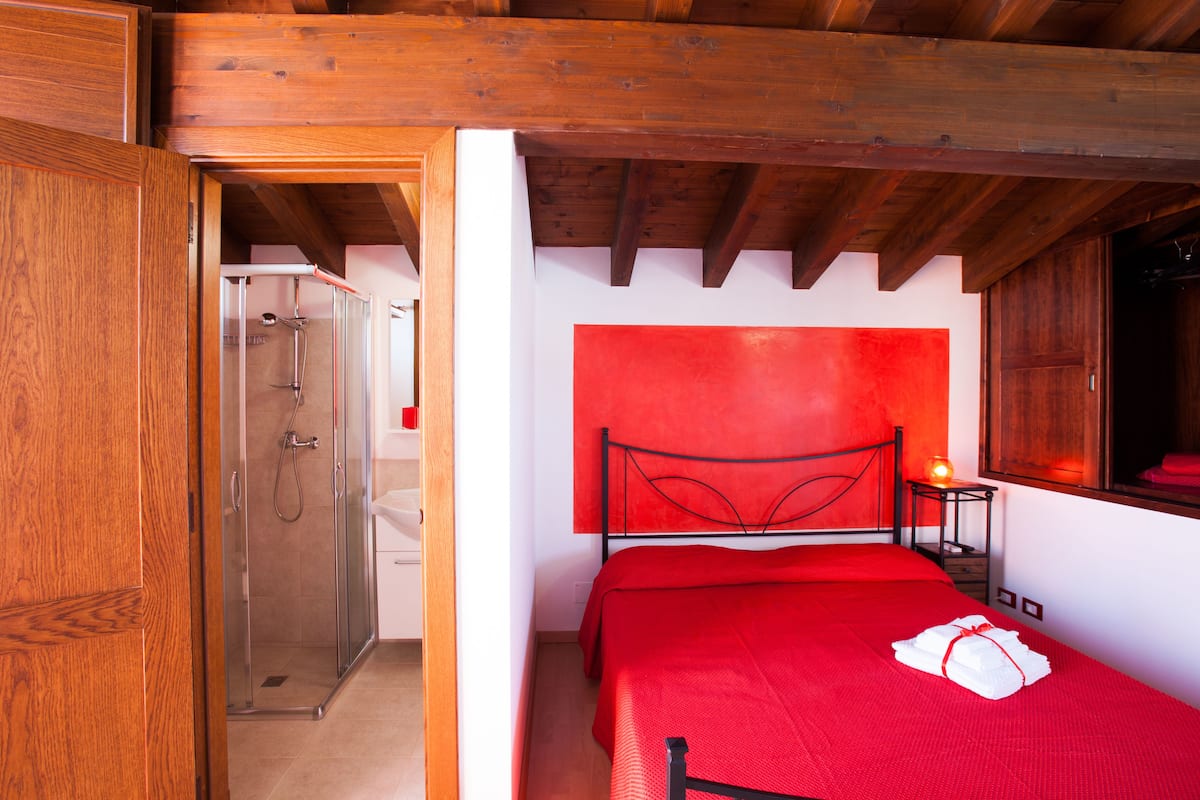 A bedroom with a red bedspread, a wooden ceiling, and a shower area.