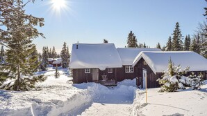 En træhytte med snedækket tag, omgivet af sneklædte træer og en klar himmel.