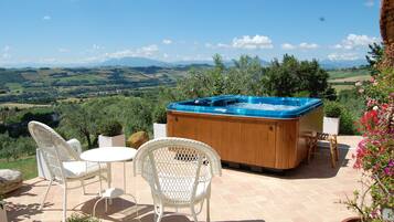 Een hottub met een houten buitenkant, omringd door witte wicker stoelen en een klein, rond tafeltje, gelegen op een terras met een prachtig uitzicht op glooiende heuvels en een heldere lucht.